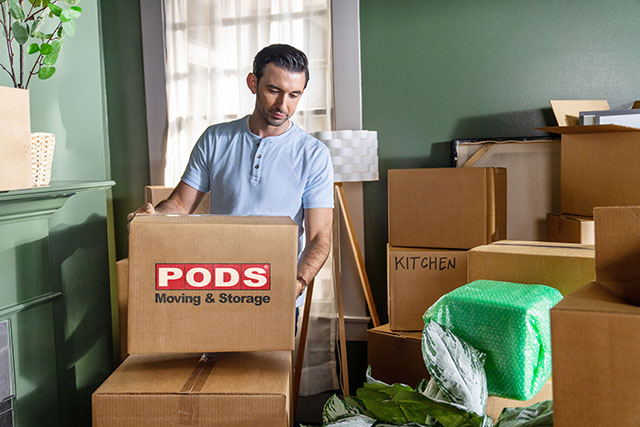A man in a living room surrounded with moving boxes as if he just moved. He is currently holding a brown box with the PODS logo displayed on it