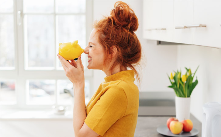A young red-haired woman in a yellow shirt is standing in her kitchen and smiling while holding a little yellow piggy bank up to her nose. The image conveys the concepts of “saving money” and an “affordable cost of living.”