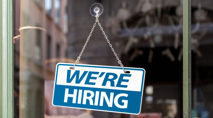 A small blue and white sign with the message “WE’RE HIRING” is hanging from a hook on the glass wall of a storefront. 