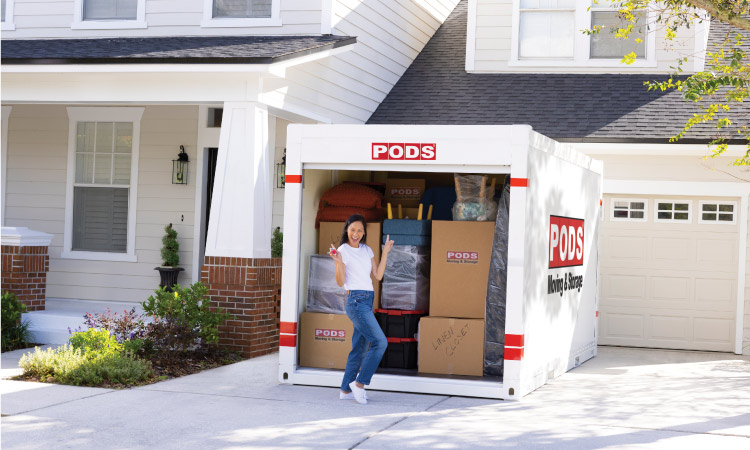 A woman is holding a key in one hand and pointing behind her with the other as she poses in front of her fully loaded PODS portable moving container in the driveway of her home. 