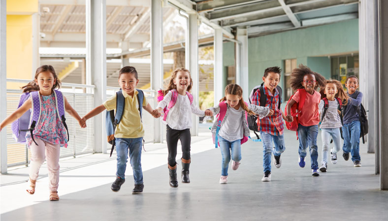 Eight happy children are wearing backpacks and running through the hallway of their elementary school while holding hands.